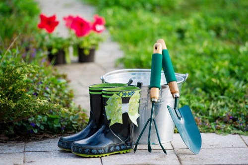 The image displays a pair of dark blue rubber gardening boots with a floral pattern on the sole, placed on a paved garden pathway. Inside and beside the boots, there are various gardening tools including a small hand rake with a wooden handle, a trowel with a wooden handle, and a metal bucket. In the background, potted flowering plants with vibrant pink blooms are arranged along a garden border, set against lush green foliage and grass. The scene appears to be in a well-maintained outdoor garden area, likely in Wimbledon, with a focus on gardening and lawn care activities typical of a residential outdoor space. The lighting suggests a bright, possibly sunny day, emphasizing the natural colours and textures of the plants, tools, and paving. Lawn Mowing Wimbledon offers gardening services in the local area, supporting garden upkeep and outdoor maintenance.