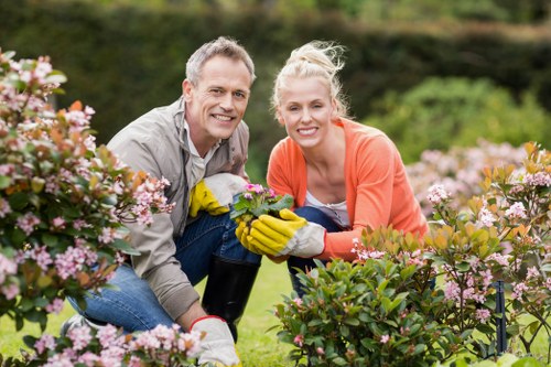 A smiling man and woman working together in a well-maintained garden, surrounded by vibrant pink and white flowering shrubs, with a lush green lawn and a background of trees and hedges. The man is wearing a grey shirt and yellow gardening gloves, holding a small gardening tool, while the woman is dressed in an orange cardigan and also wearing gloves, engaging with the plants. The garden features neatly trimmed bushes, a spacious grassy area, and a natural outdoor setting typical of residential landscaping in Wimbledon, with soft daylight enhancing the natural colours and textures of the garden; this scene exemplifies professional gardening and lawn care services provided by Lawn Mowing Wimbledon in the local area.