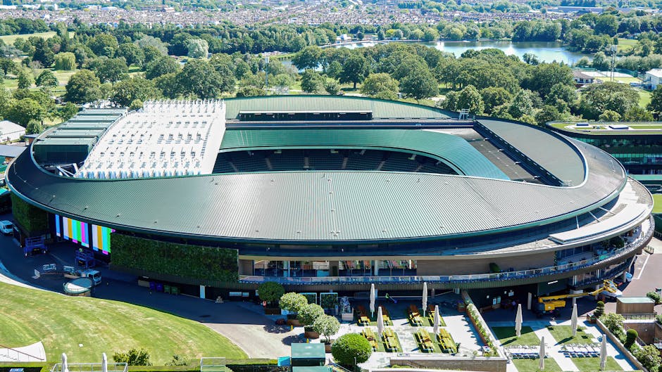 An aerial view of a modern, oval-shaped sports stadium with a green metal roof featuring a central open section. Surrounding the stadium, there are neatly maintained garden beds, pathways, and outdoor